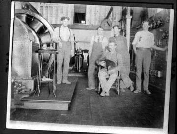 Five men (one holding a dog) standing next to the switchboard and generators in Mill Creek #1 Hydro Plant