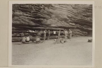 Camp on right bank at Mile 115.  Seymour and Jordan seated left, Joe Desloge kneeling, MacDonald and Marston center, Sanderson in back of groceries, Taylor and Desloge at extreme right