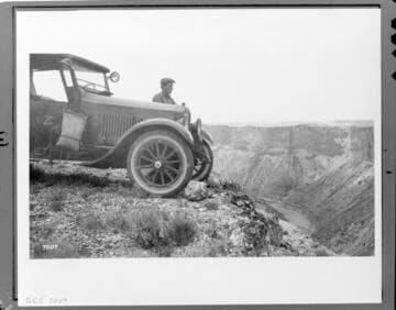 Man standing by 1920's automobile with water bag hanging on door at the edge of what is probably the Grand Canyon