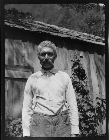 An unidentified Native American man standing in front of wooden house