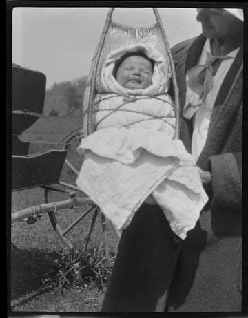 A Native American baby in a carrier or cradle-board, being held by unidentified woman